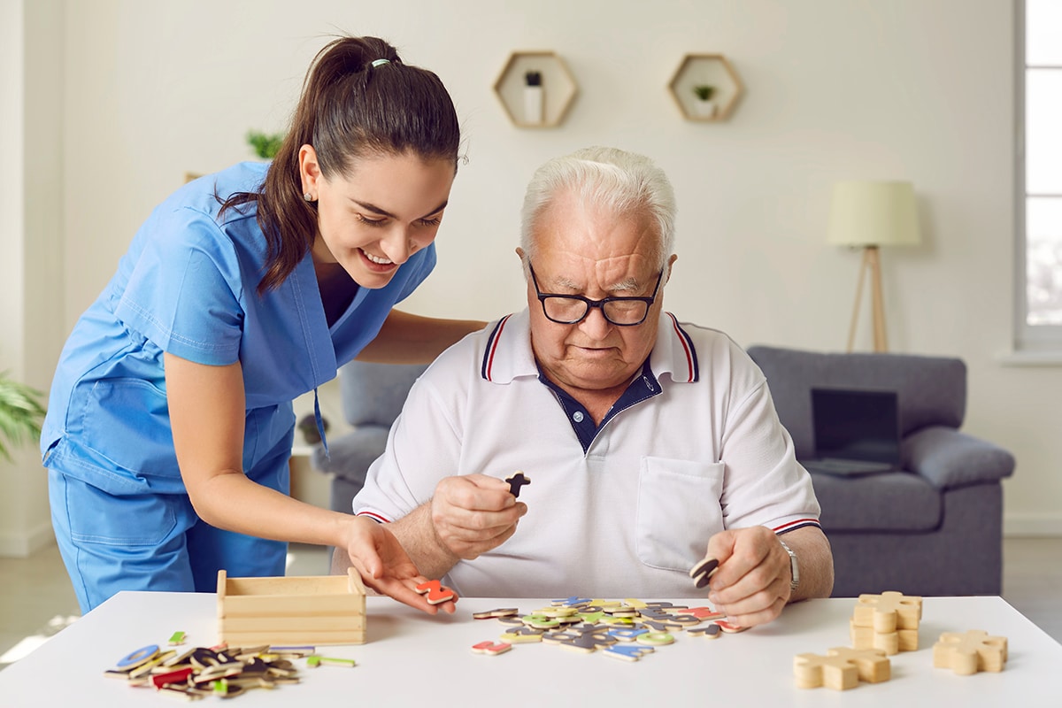 Nurse in geriatric clinic or retirement home helping patient with alphabet puzzle. Senior man sitting at desk and playing games with letters. Old age, dementia, Alzheimer's disease, therapy concept